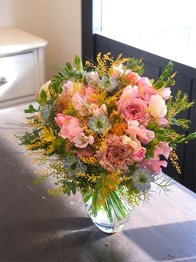 定年退職される女性に、感謝とお祝いの気持ちを込めた贈り花です。フリージア‘エアリーピーチ’、ラナンキュラス‘リナ ノエル’、チューリップ‘ピューリッツァー’、スイートピー‘たきひめ’と、ニュアンスの異なるピンクの花を集め、ニゲラ‘ミスジーキル’の水色と金葉アカシア‘つとむ’の黄色で華やかなアレンジを制作しました。
