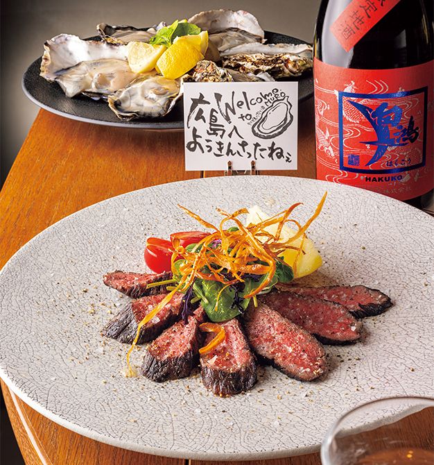 奥は「かきむすめ」（１個770 円）。呉沖で育ち、海流の上下運 動により殻が深く、身も詰まって いる。手前は「比婆牛のモモ 檜 焼きステーキ」（100㌘4600 円）。 呉・盛川酒造の「白鴻」とともに。 鯉ラベルは広島限定。