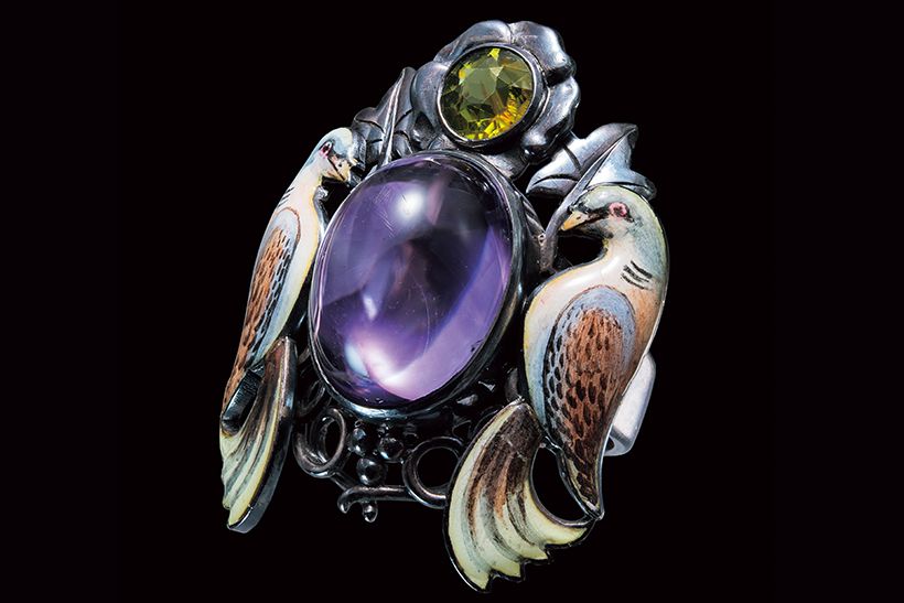 英国で1920年頃制作された《アメシストと二羽の鳥》。作者のジョージ・ハントは、ウィリアム・モリスを中心としたアーツ＆クラフツ運動の影響を受けた英国のジュエラー。カボションのアメシストとペリドット、鳩のエナメル装飾からなる芸術的な一品。