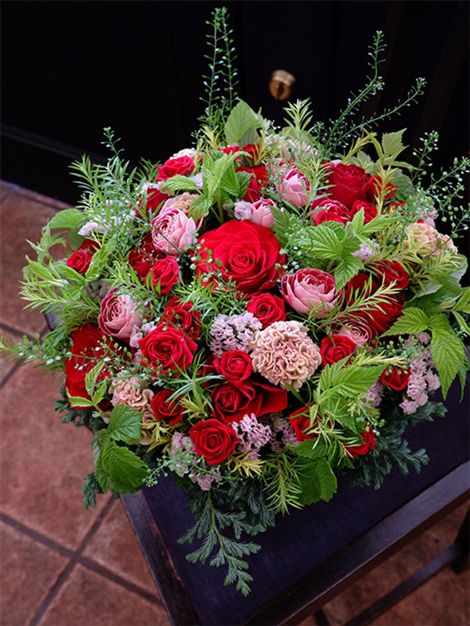 妹さんの還暦祝いに、お姉さんからの贈り花です。還暦（60歳）といえば、テーマカラーは赤ということで、鮮やかな赤バラ‘ブリランテ’をメインにしたアレンジを制作しました。あでやかなピンクのバラ‘チアフル’、ページュがかったピンクのケイトウ‘プティフルーツ フェアリーローズ’、透明感のあるピンクのスターチス‘ソロモン’と、ニュアンスが異なるピンクを脇役として利用しました。ほかにバラ‘レディーラブ’、メラレウカ‘レボリューションゴールド’、コニファー‘ブールヴァール’、フランボワーズなどを使用。