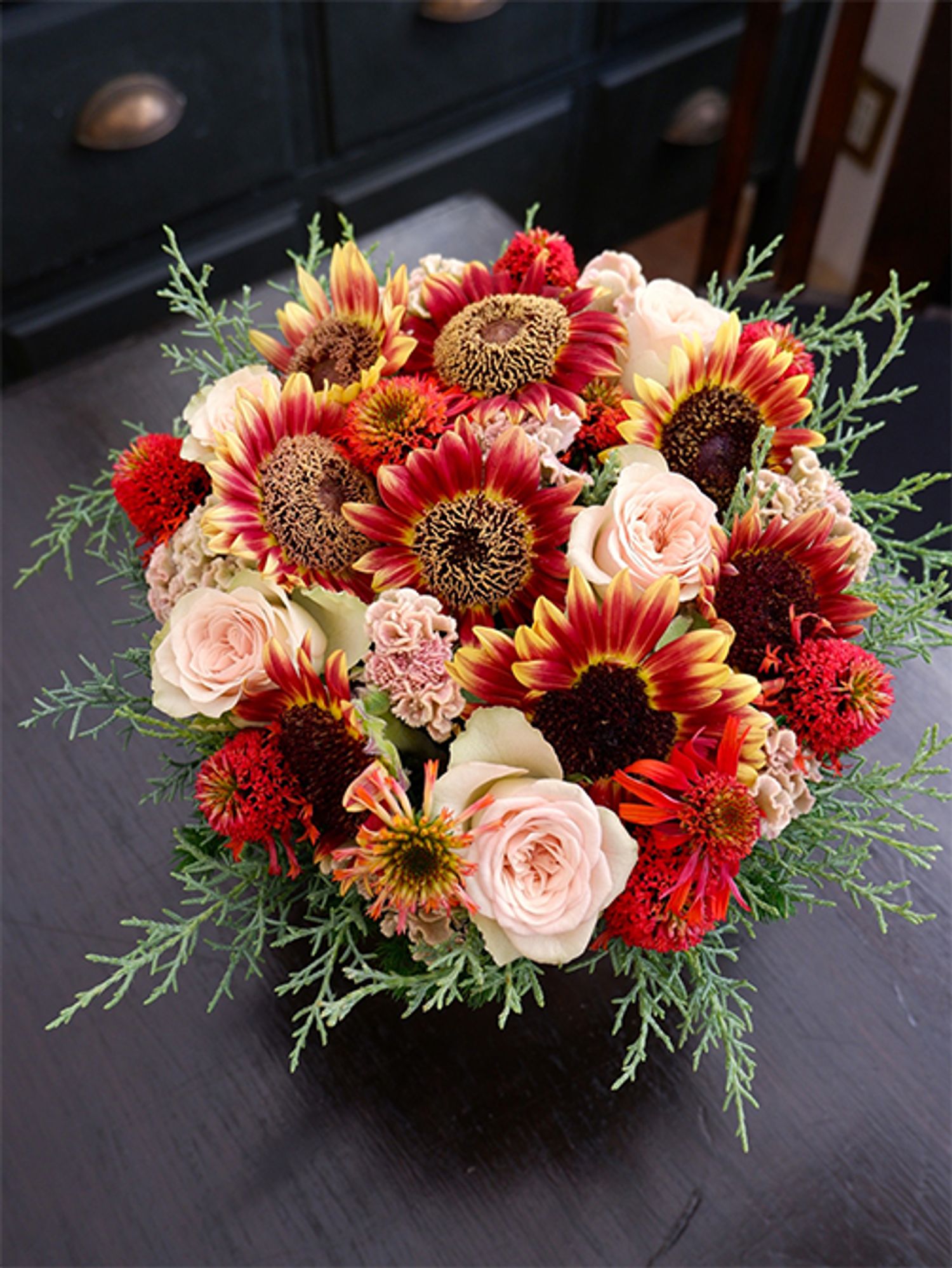 お誕生日の贈り花に、ケイトウ‘プティフルーツ フェアリーローズ’を秋色のヒマワリと合わせたアレンジを制作。個性の強い赤系の花をケイトウの落ち着いたピンクがバランスよくまとめてくれます。秋色のヒマワリは‘フロリスタン’。ほかにバラ‘パンテオン’、エキナセア‘レイラ’を使い、コニファー‘ブルーアイス’をアウトラインにあしらいました。
