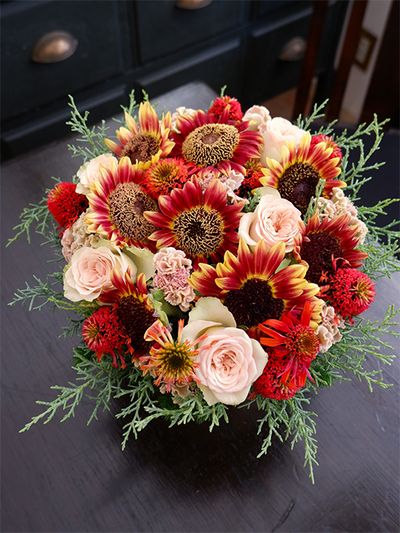 お誕生日の贈り花に、ケイトウ‘プティフルーツ フェアリーローズ’を秋色のヒマワリと合わせたアレンジを制作。個性の強い赤系の花をケイトウの落ち着いたピンクがバランスよくまとめてくれます。秋色のヒマワリは‘フロリスタン’。ほかにバラ‘パンテオン’、エキナセア‘レイラ’を使い、コニファー‘ブルーアイス’をアウトラインにあしらいました。