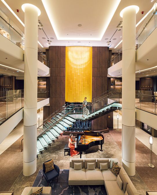 レセプション前の3層吹き抜けのロビーでは、人間国宝の漆芸家・室瀬和美氏の高さ約9メートルにも及ぶ壮大な漆芸作品が迎える。海面に降り注ぐ光の情景が表現されたこの作品は、蒔絵の技法で金箔と螺鈿があしらわれている。 ドレス66万円　バッグ87万100円　靴23万1000円／すべてブルネロ クチネリ（ブルネロ クチネリ ジャパン）