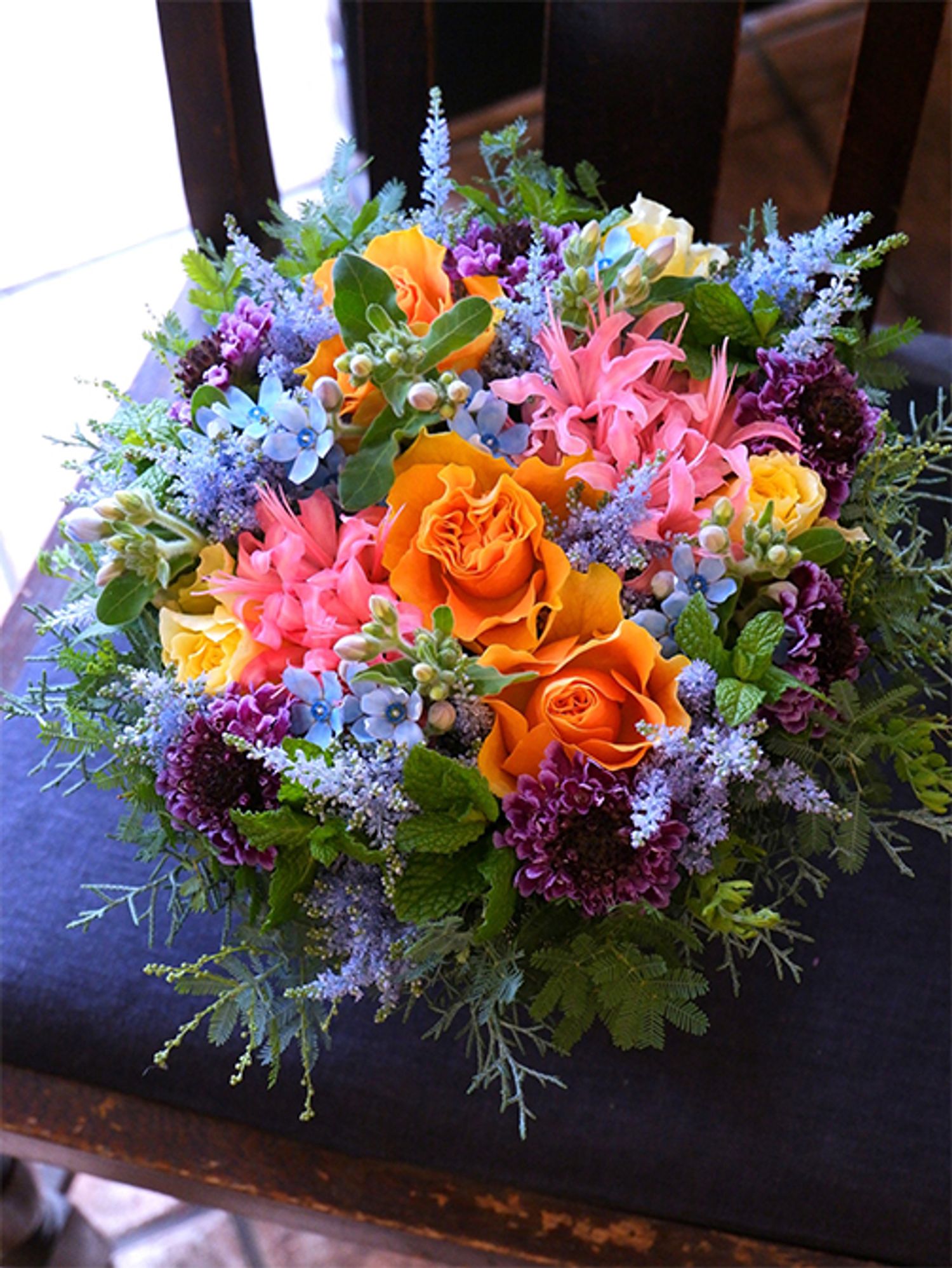 バラの黄色とオレンジ色、ダイアモンドリリーのピンク、スカビオサのパープル。華やかな多色合わせのアレンジでひときわ存在感を放つのがオキシペタラム‘ピュアブルー’の水色とくっきりした星の花形です。バラは‘スフィンクスプレゴ’と‘レモンフォセット’、スカビオサは‘カーブアウトフェイト’を使用。染めのアスチルベ‘ライトブルー’でブルーをボリュームアップしました。ほかにスペアミント、銀葉アカシアなどを使用。高知県で暮らすお祖母様へ、東京のお孫さんたちから誕生日の贈り花です。