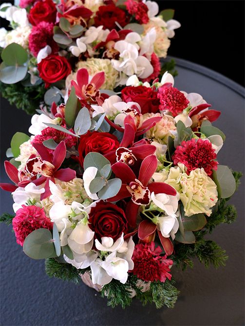 京都と大阪、2カ所へのお歳暮のオーダーをいただき、お揃いで制作しました。高級感や特別感を演出したいときにおすすめなのが、赤バラとランの組み合わせです。このアレンジでは、バラ‘キララ’とシンビジウム‘ミミ ルシファー’を合わせました。スカビオサ‘プリエルージュ’の透明感ある赤を足して、赤のグラデーションの幅を広げています。カーネーション‘モチ’、宿根スイートピーの白花、シルバーリーフのユーカリを足して、全体を明るい印象に。