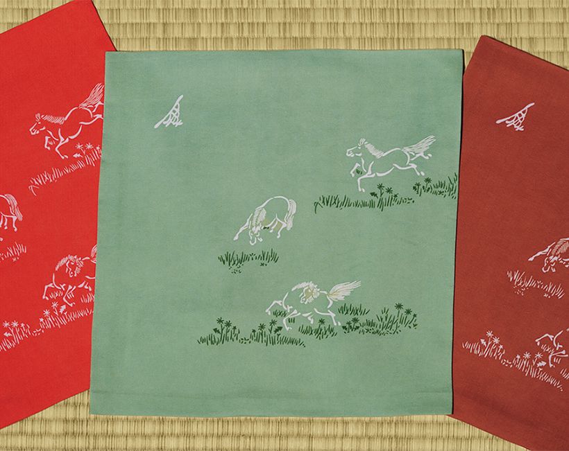 家元が干支にちなんでデザインした令和8年の袱紗。草木が生い茂る中に躍動感溢れる3頭の馬を配した意匠「勇馬文」には、新年の飛躍を願う心が込められている。左から曙紅、天青、檜皮色の3色。