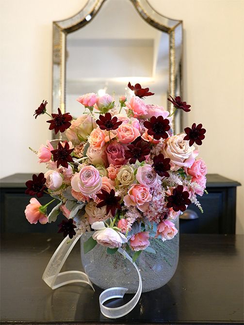 バラやラナンキュラスなどピンクの花を束ね、少し飛び出すようにチョコレートコスモスをあしらったウエディングブーケ。チョコレートコスモスのシックで濃い花色が加わると、かわいらしいピンクが少し大人っぽい表情に。バラは‘ウエストミンスターアビー’、‘アヴニール＋’、‘デザート’、‘リリカ’と、ニュアンスの異なるピンク4種を集めました。ほかにラナンキュラスの‘エルフード’と‘ポワント’、アスチルベ‘ライトピンク’、ダスティーミラーなどを使用。