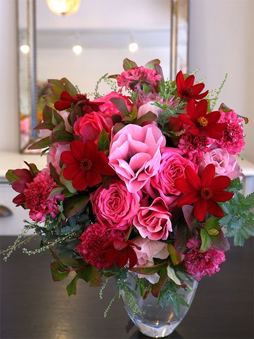 寒い時期にぬくもりを感じさせる赤×ピンクのブーケを。赤はチョコレートコスモス‘ノエルレッド’、濃淡ピンクはスカビオサ‘プリエルージュ’、バラの‘アヴニール＋’と‘ローズシノ’。ギンヨウアカシア、ヒペリカムなどのリーフをあしらって仕上げました。チョコレートコスモスの赤は、ボリューム感のあるバラやスカビオサに負けないほどインパクトがあります。