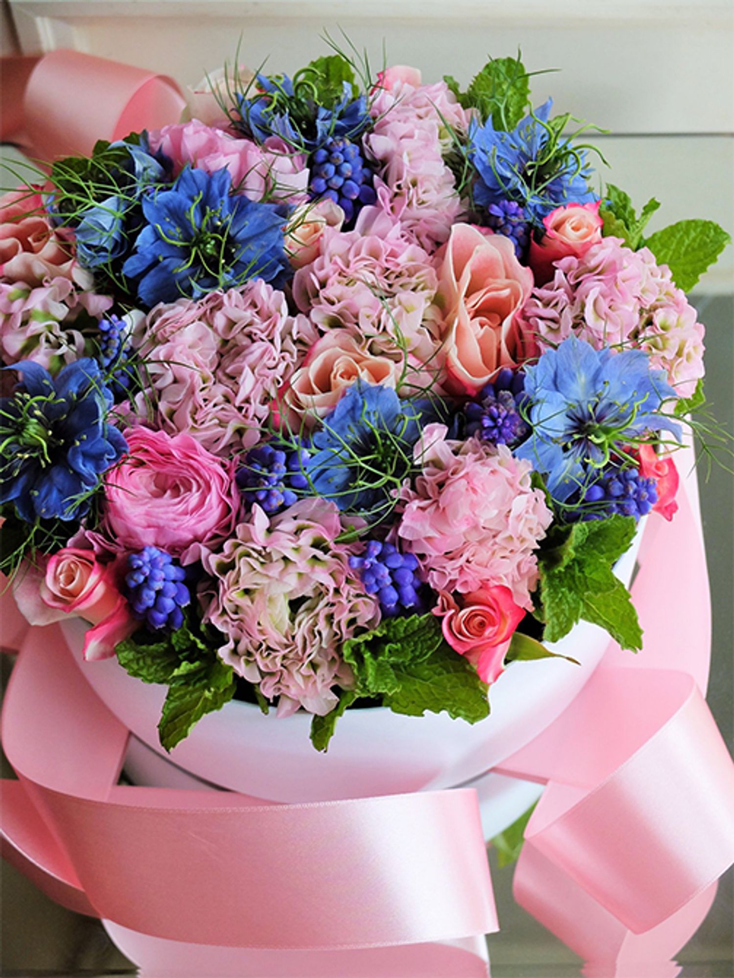 春のピンクとブルーで華やかな花箱を作成しました。ピンクはラナンキュラス‘ポンポン ハーマイオニー‘とバラ‘ストロベリーモンローウォーク’。ブルーはムスカリとニゲラ‘ミスジーキル’。スペアミントのフレッシュグリーンを添えて仕上げました。ピンク×ブルーの花色合わせは本当にエレガント。ピンクだけではかわいすぎるかなと感じたときにおすすめです。直径20cmのボックスを使用。