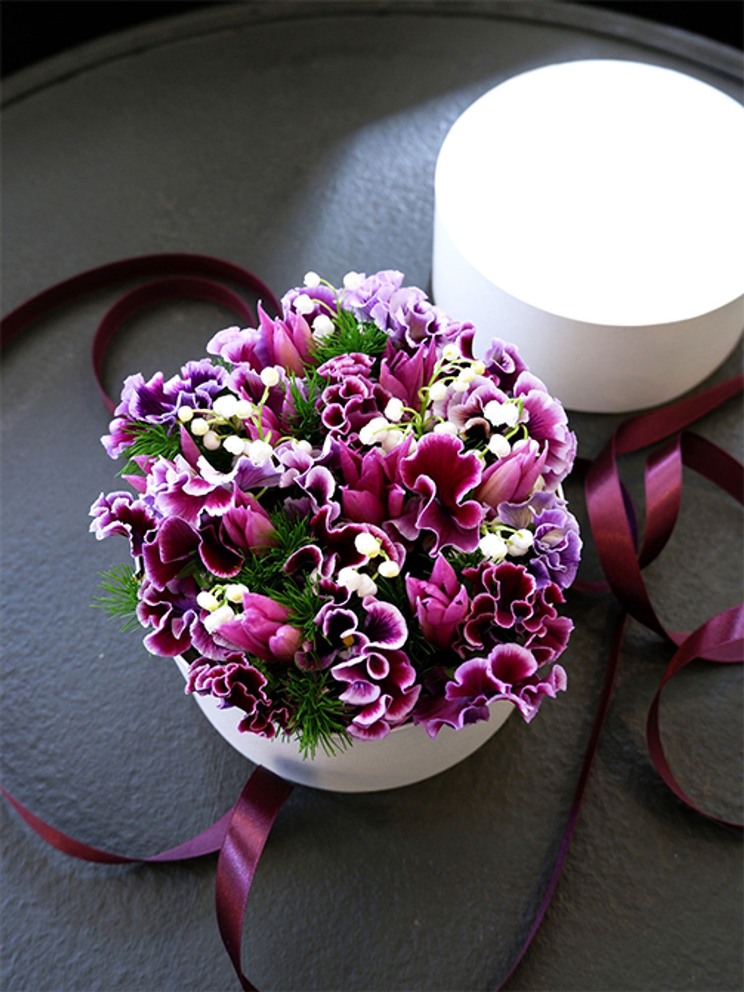 花箱に利用しやすいスズランが出回る時期です。紫色のパンジー‘フリズルシズル バーガンディー’とチューリップ‘夢の紫’を合わせて丸いボックスに詰めました。箱の直径は15cm。小さな箱からあふれる春の紫色がエレガントな雰囲気です。