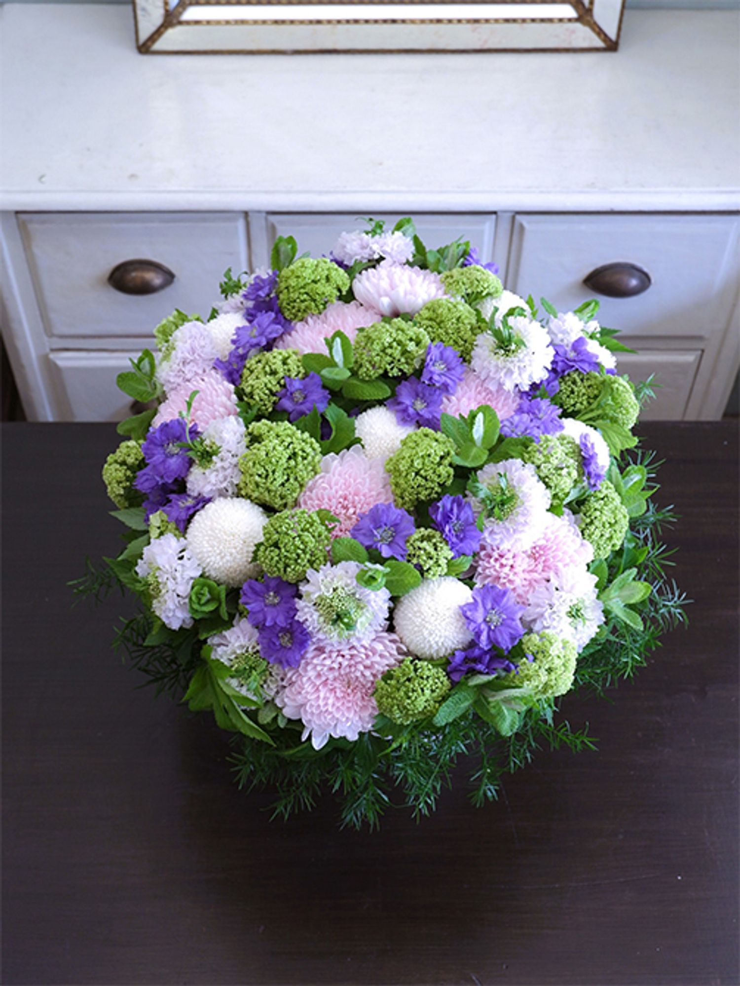 白花の‘ちょんまげ トノ’を使い、涼しげな花色合わせのアレンジを制作。マム‘セイ ヌーベル’とピンポンマム、デルフィニウム‘ラベンダーサファイア’、ビバーナム・スノーボール、アップルミントなどを使用。スカビオサの花心からちょこんと飛び出る葉っぱが楽しさを引き出します。