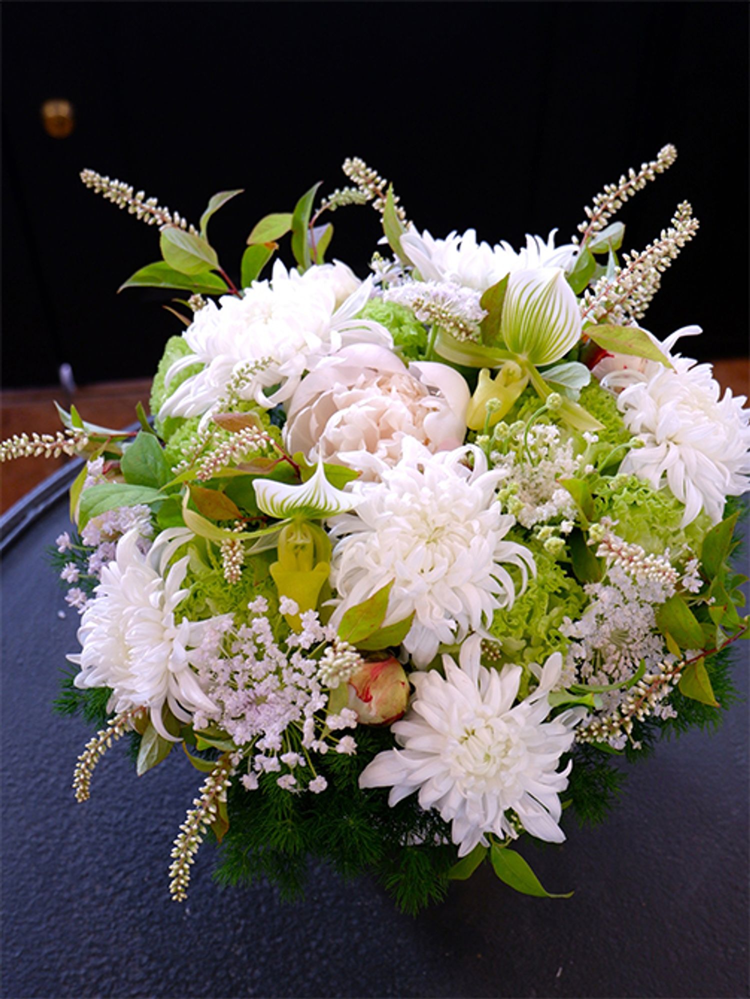 ヒメリョウブは白といっても少しクリームがかった花色なのですが、マムの白花と合わせると、そのニュアンスの違いがよくわかります。明るさのある純白の花の中で、周囲に散らしたヒメリョウブは引き締め役にもなっています。マムは‘セイ マノアホワイト’を使用。ほかに、シャクヤク‘ホワイトピーチ’、リシアンサス‘アンバーダブルモヒート’、パフィオペディルム、ブルーレースフラワー‘ピュアレディ’を選びました。