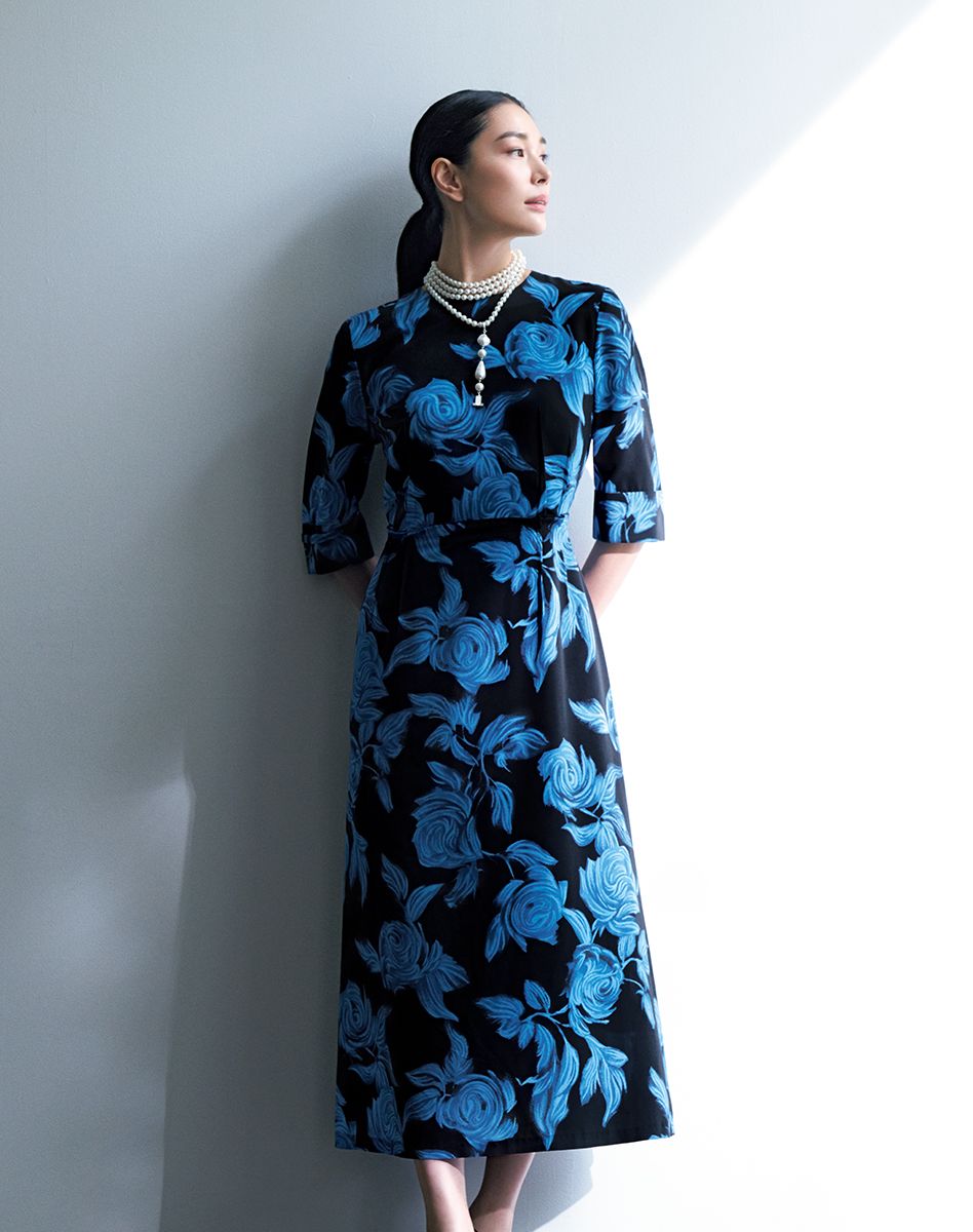 漆黒に咲き誇る大輪の青いバラが、凜とした佇まいを印象づけるロングワンピース。素材は国内有数の北陸産デシンで、パウダーをはたいたようなまろやかな風合いが、成熟した女性にふさわしい品格を醸します。ウエストは、あえて表に配した立体的なダーツが高めの位置で引き締め、すらりとした美しいスタイルを演出。くるみボタンをあしらったカフスも、ひじを包む優雅な袖丈と相まって、洗練された余韻を残します。ワンピース16万5000円 ネックレス4万9500円/ともにランバン コレクション